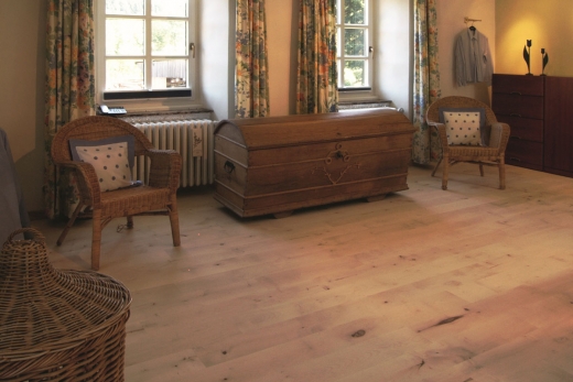Et rum med lyse gulvbrædder i massiv ahorn. To vinduer med blomstrede gardiner lukker dagslys ind. Der er to kurvelænestole og en gammel trækiste i rummet. Gulvbrædderne har en naturlig, lys farve.